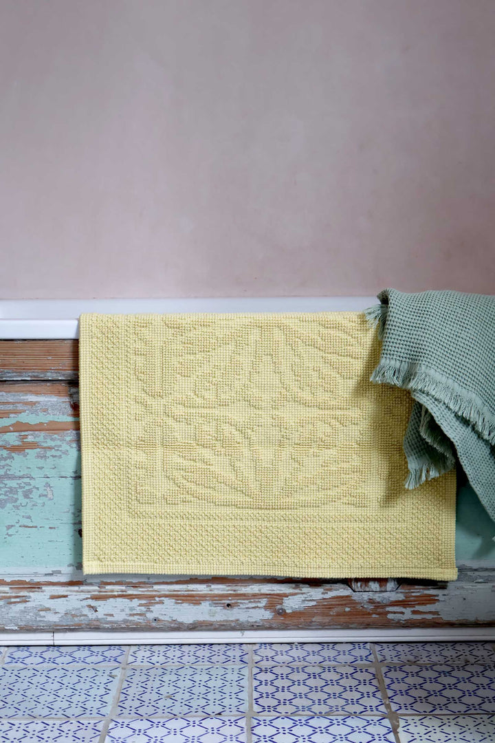 Yellow textured bath mat draped over a wooden surface with a pink wall in the background
