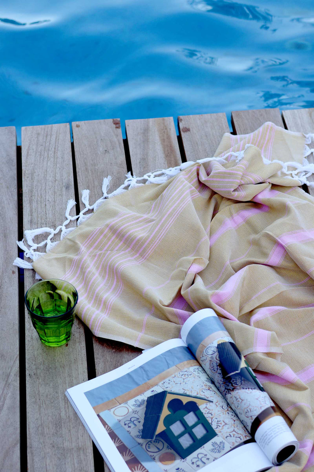 Beige towel with pink accents on a wooden deck by a poolside, with a green glass and rolled-up magazine.
