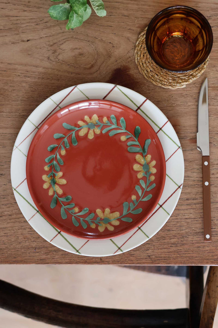 A ceramic dinner plate set on a wooden table, featuring a red-orange plate with a green and yellow floral wreath design, layered over a white charger plate with a green and red plaid border. A brown-handled knife and an amber glass tumbler are placed nearby.