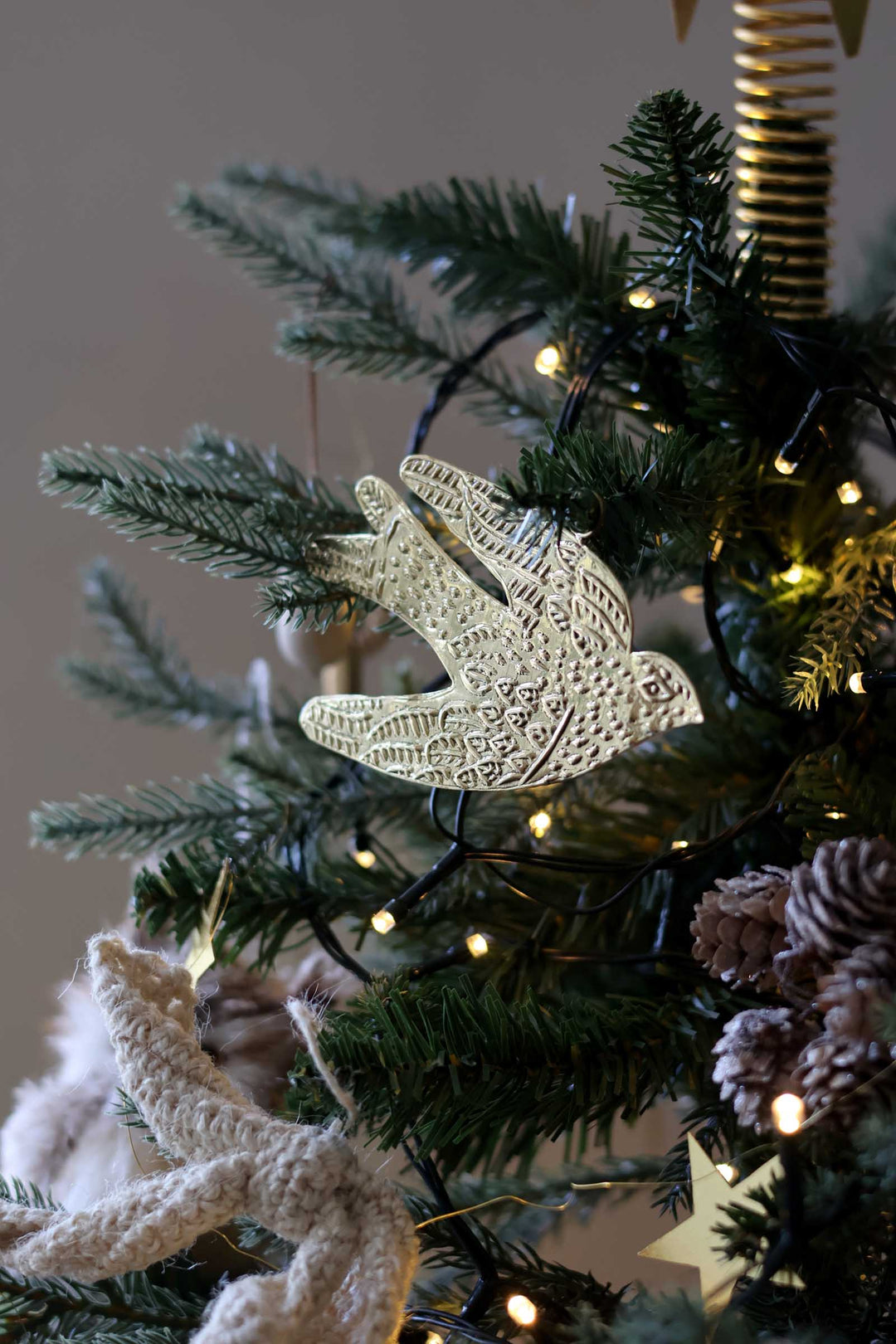 A close-up of a decorated Christmas tree branch with a carved white bird ornament hanging among green pine needles, warm string lights, and rustic natural accents.