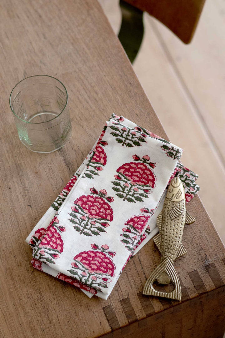 A folded white cotton kitchen towel with a pink and green floral block-print pattern, styled on a rustic wooden table next to a clear glass tumbler and a decorative metal fish bottle opener.