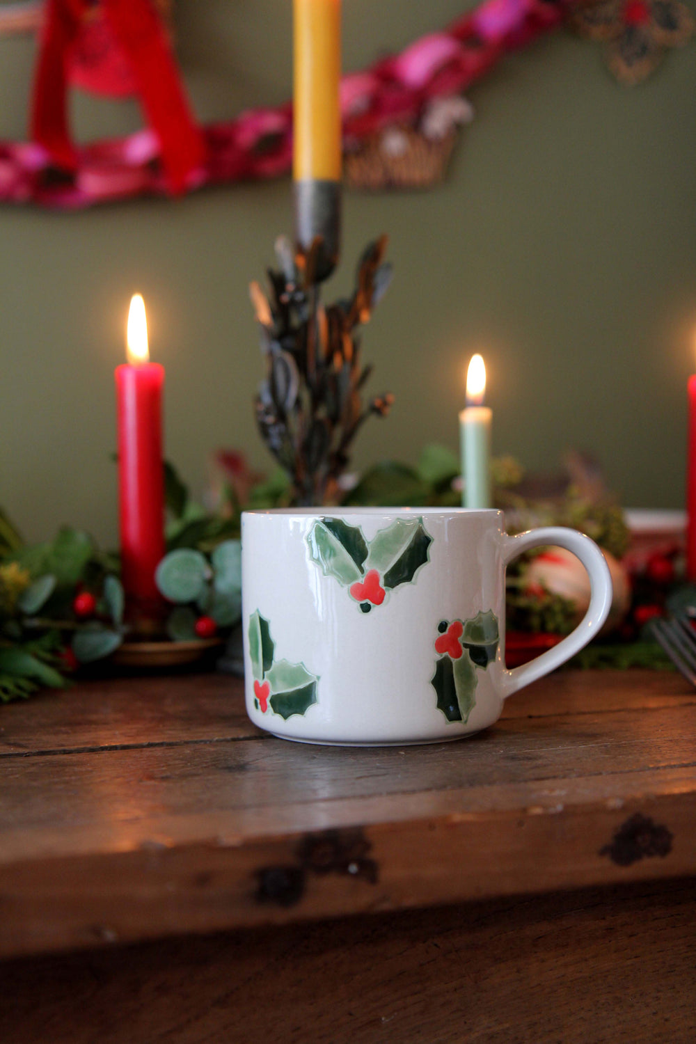 White mug with holly design on a wooden surface with candles and greenery in the background
