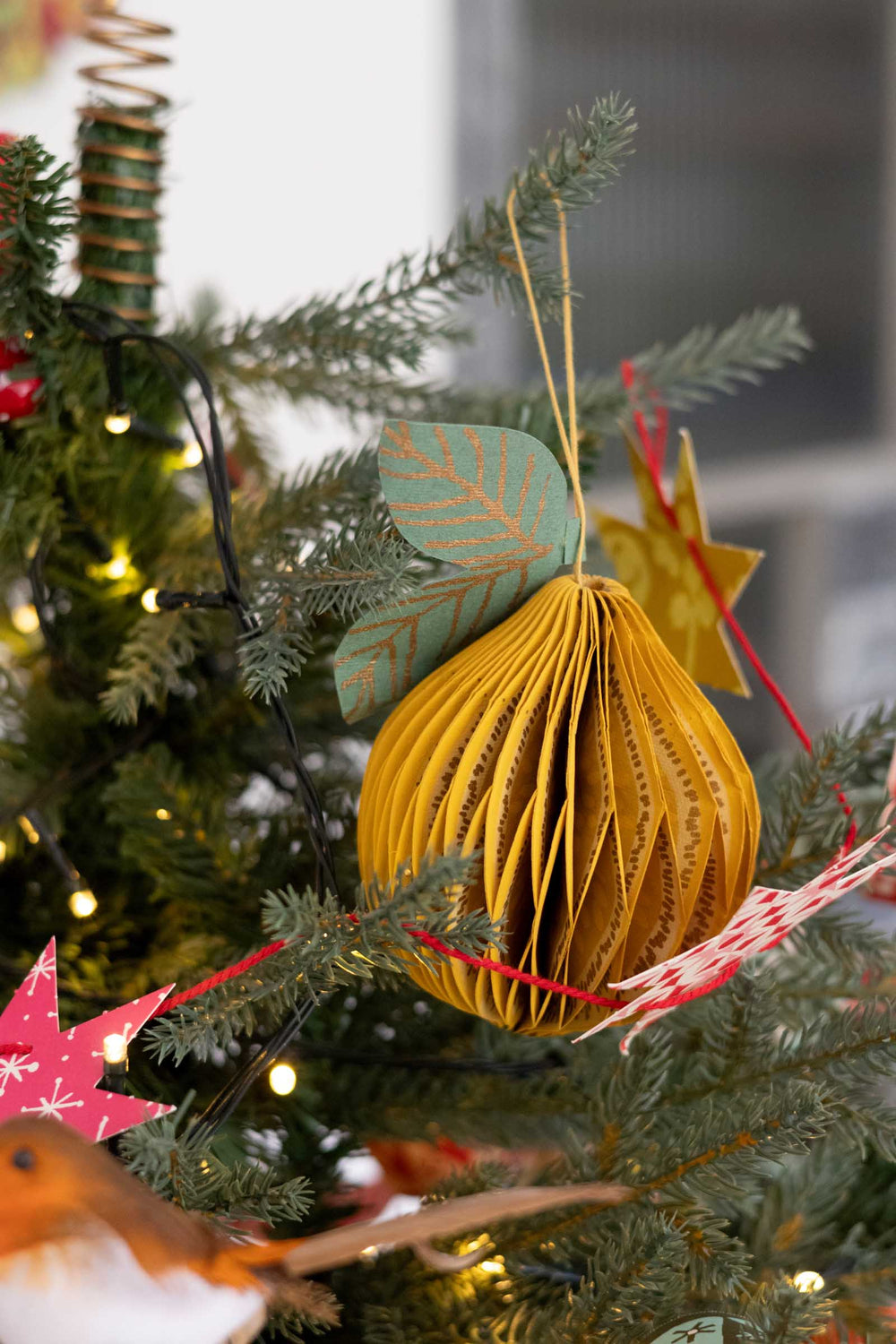 A yellow paper accordion-style Christmas ornament hanging on a decorated evergreen tree with warm string lights and red ribbon accents.