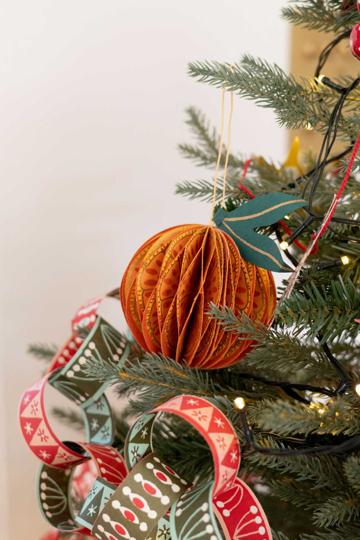 An orange ribbed paper ball Christmas ornament hanging on a green Christmas tree, surrounded by patterned red and green ribbons and small warm string lights.