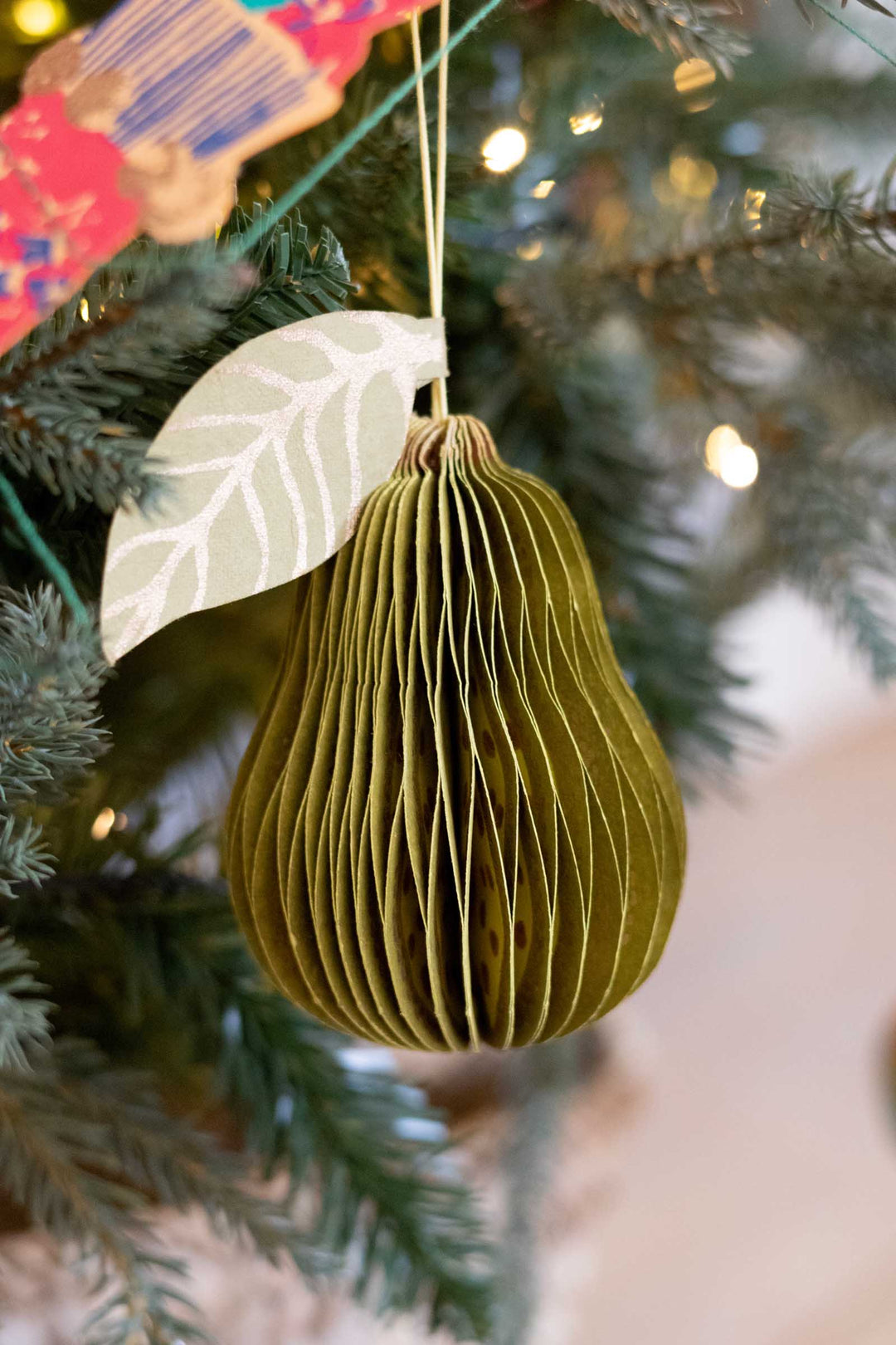 A green pear-shaped hanging ornament made of pleated paper, decorated with a white leaf accent, hanging from a Christmas tree with warm lights in the background.