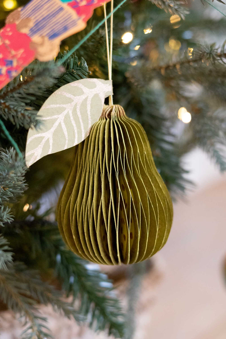 A green pear-shaped hanging ornament made of pleated paper, decorated with a white leaf accent, hanging from a Christmas tree with warm lights in the background.