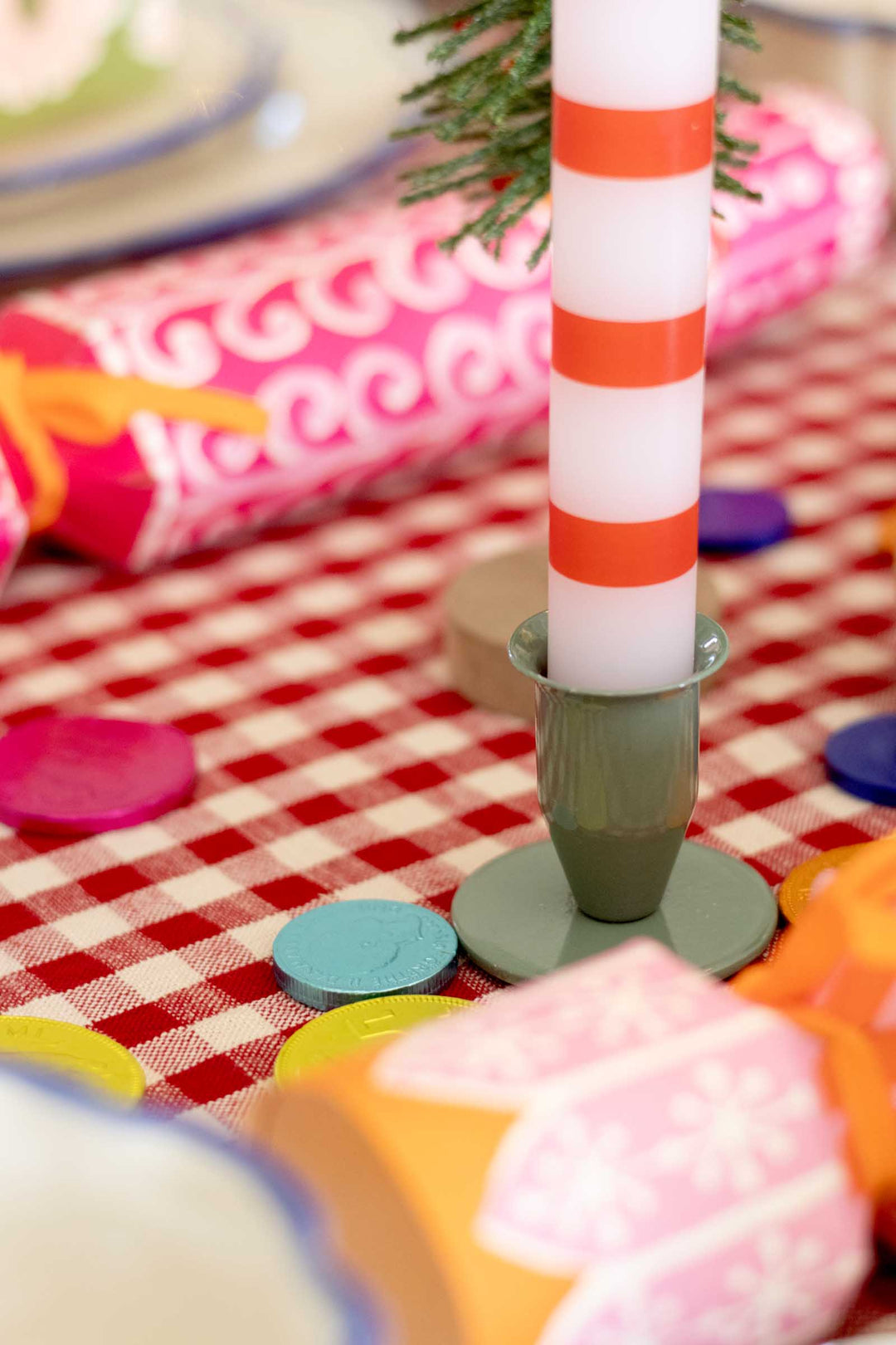 A close-up of a white candle with orange stripes in a gray-green holder, set on a red-and-white checkered tablecloth with colorful confetti and wrapped gifts in the background.