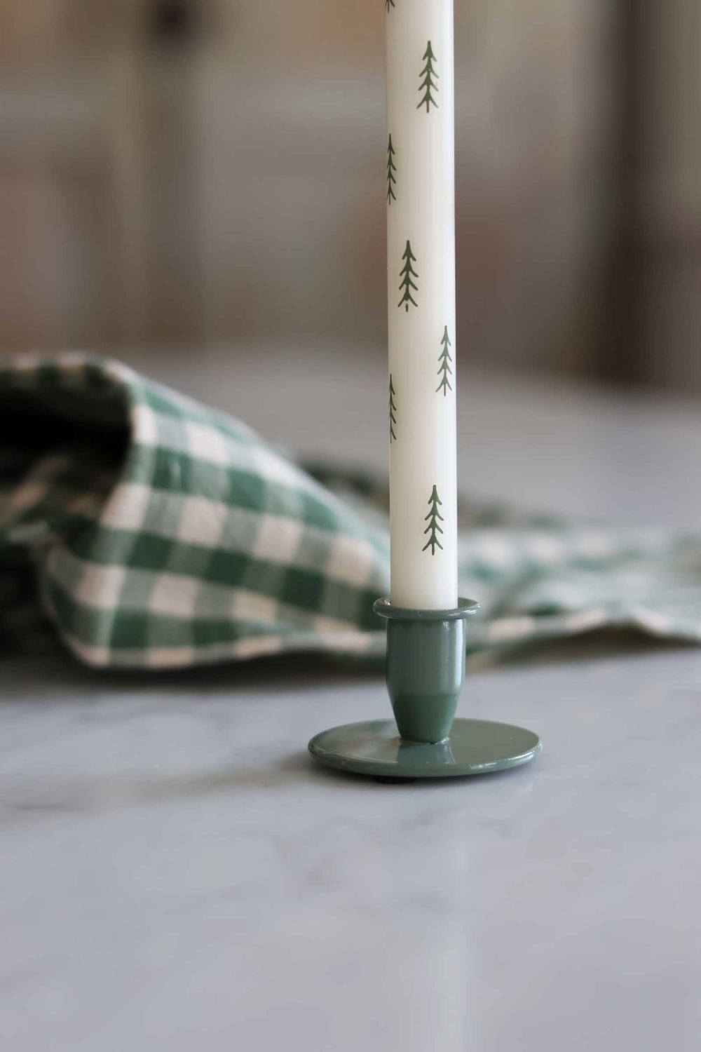 A white taper candle with a green pine tree pattern, displayed in a sage green metal candlestick holder on a light marble surface with a green and white checkered cloth in the background.