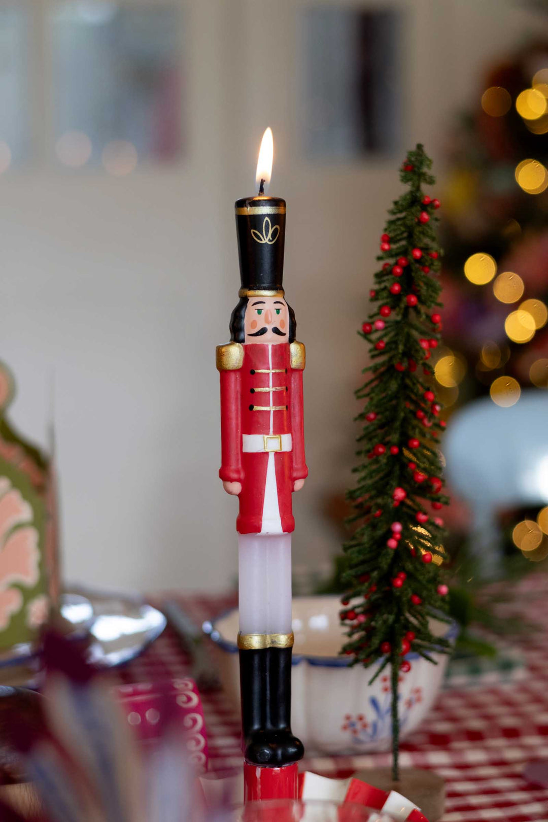 A lit Christmas nutcracker candle with a red and black soldier design, standing on a table next to a small decorated Christmas tree with warm blurred lights in the background.