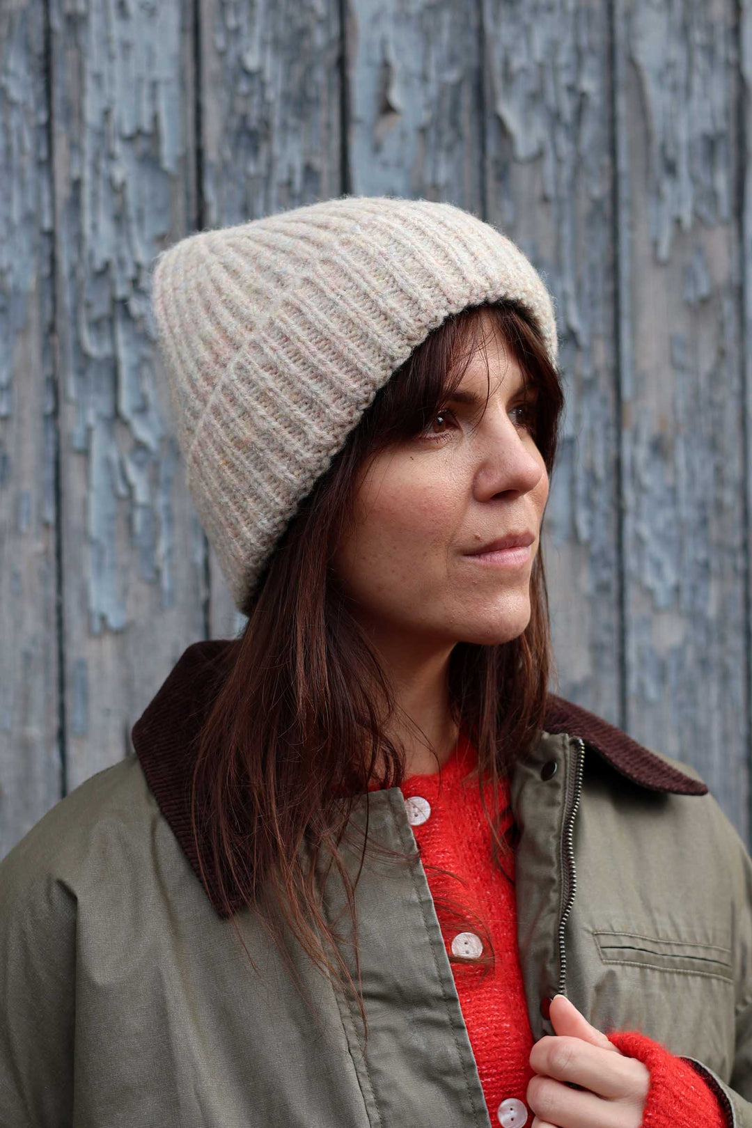 A woman wearing a beige ribbed knit beanie hat, an olive green jacket with a brown corduroy collar, and a red knit top, standing in front of a weathered blue-gray wooden wall.
