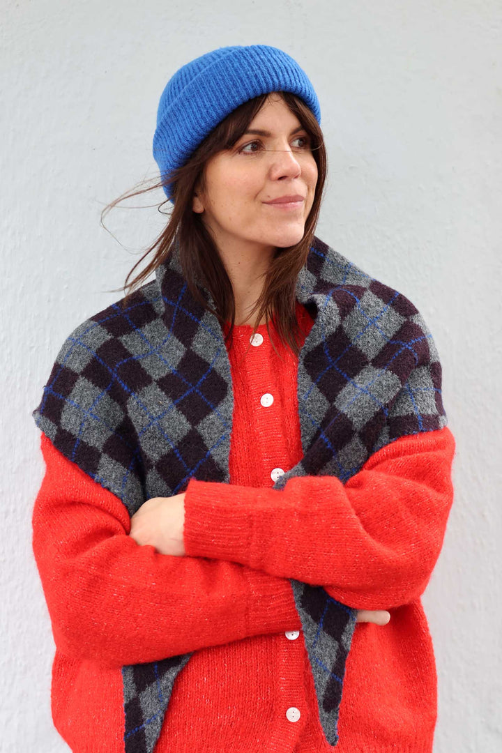 A woman wearing a bright blue knit beanie, a red knit cardigan, and a dark gray and navy argyle wool wrap shawl, standing against a light gray wall.