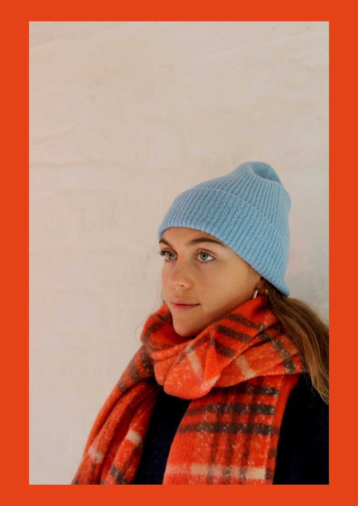 Person wearing a blue knit beanie and orange plaid scarf against a beige background