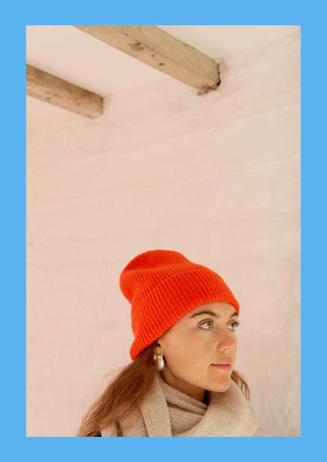 Person wearing a bright orange knit beanie against a light pink wall.