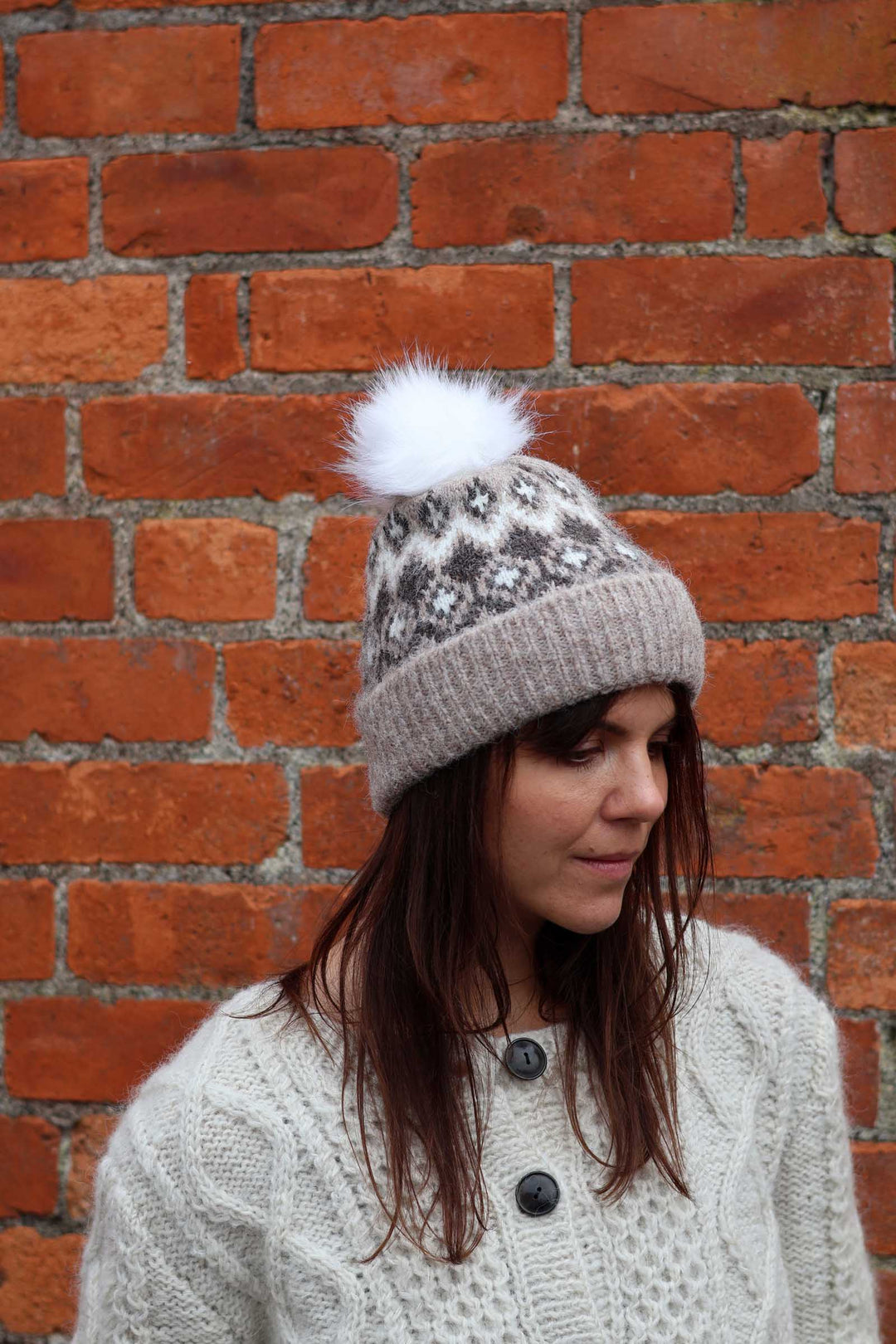 A woman wearing a gray knit beanie with a white and gray patterned snowflake design and a fluffy white pom-pom, standing in front of a red brick wall.