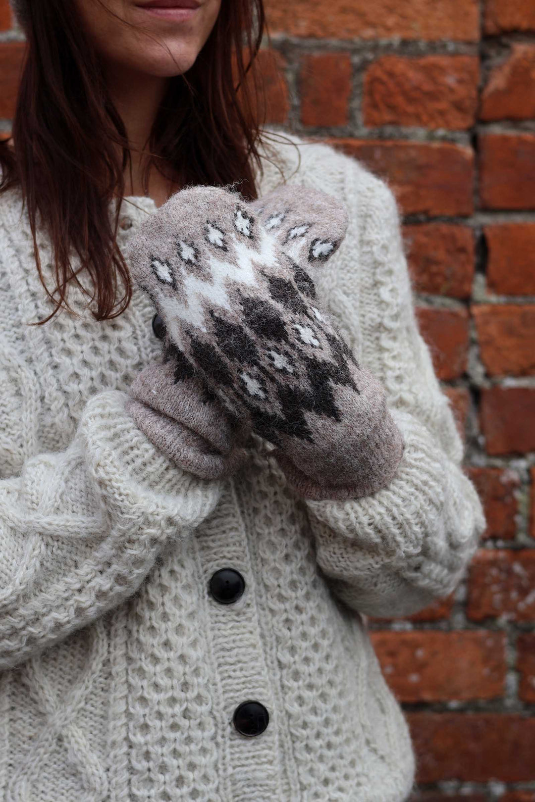 A close-up of a person wearing cream cable-knit cardigan sweater and knit mittens with a gray and white geometric pattern, standing in front of a red brick wall.