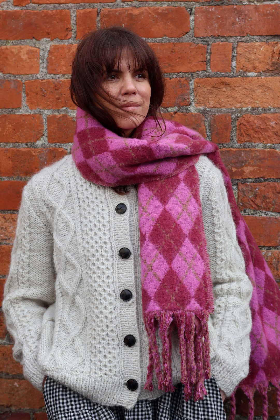 A woman wearing a chunky cream cable-knit cardigan and a thick pink and magenta argyle patterned scarf, standing against a red brick wall.