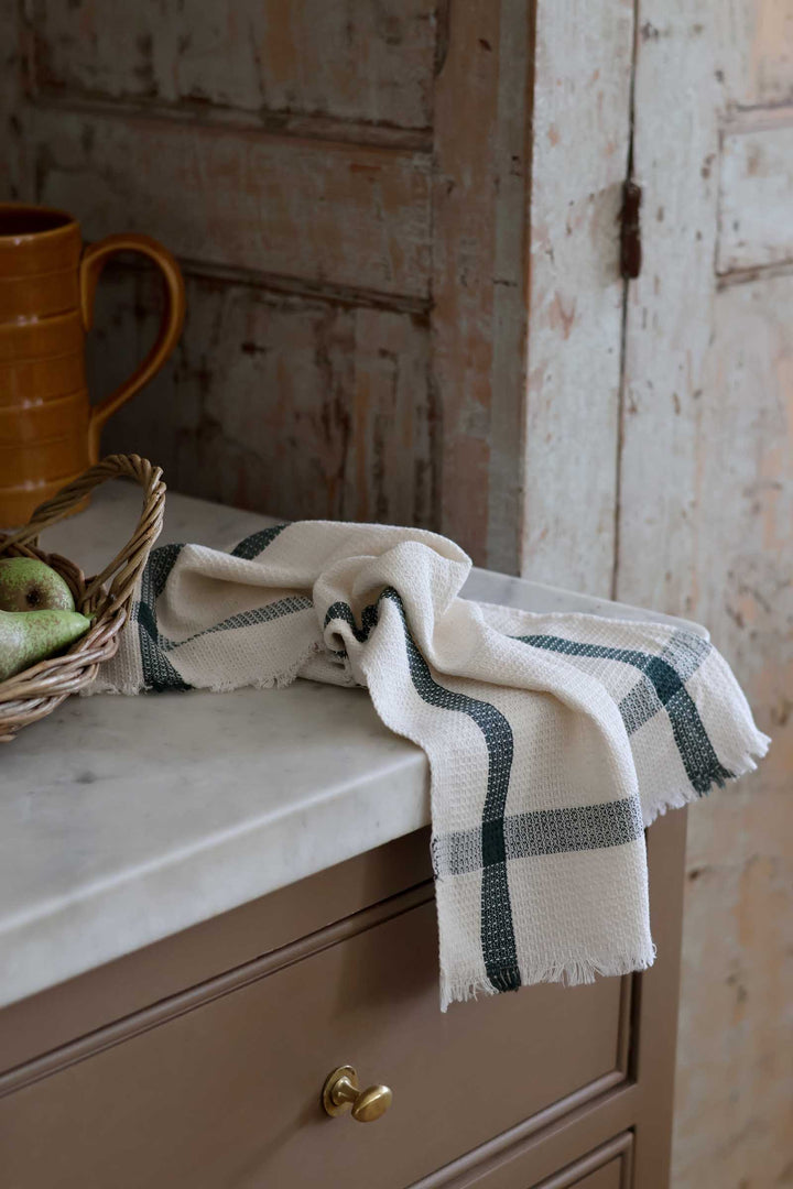 A cream-colored woven kitchen towel with dark green plaid stripes and fringed edges, draped over a brown wooden cabinet next to a wicker basket with green apples and a rustic amber pitcher.