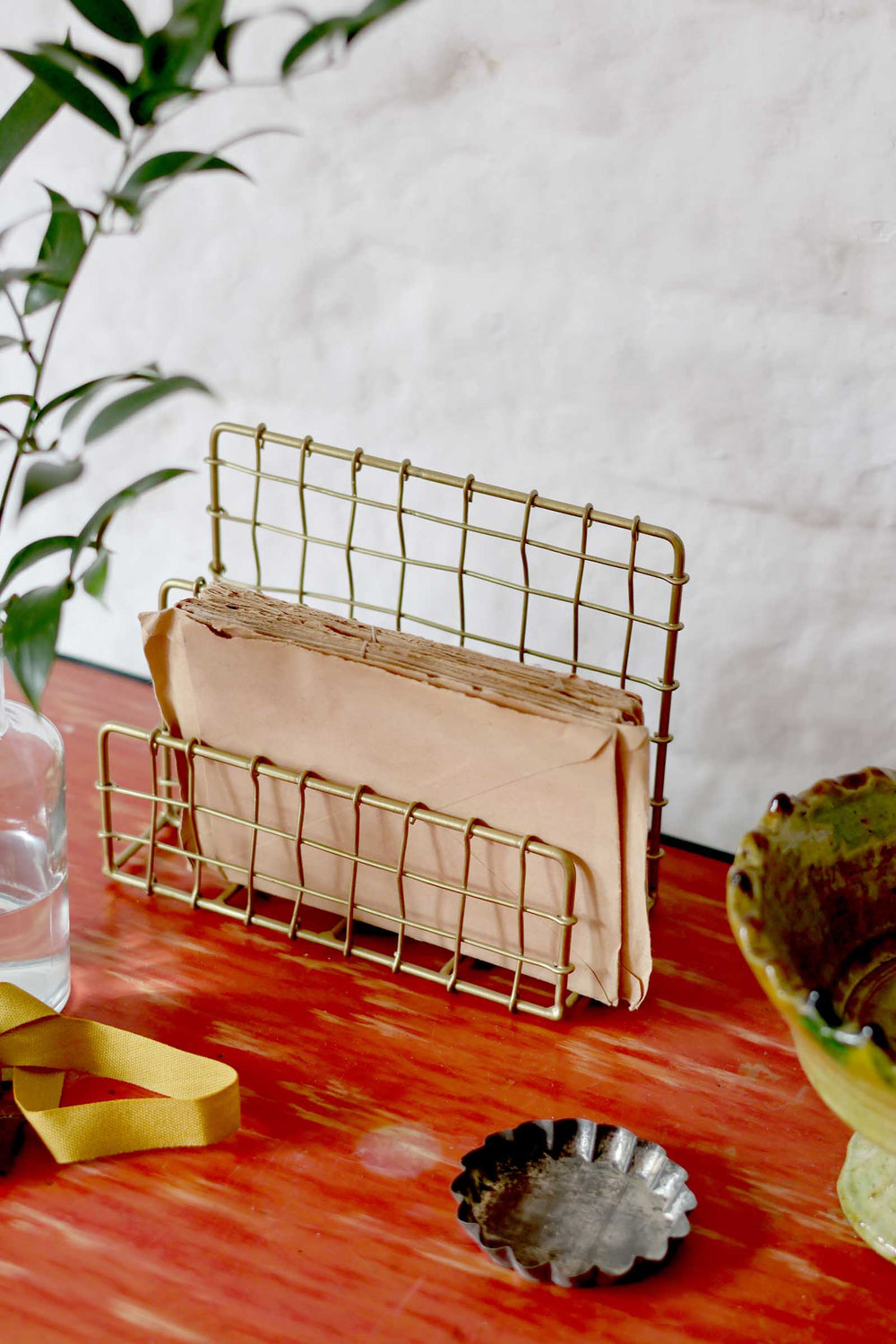 Gold wire letter holder with beige envelopes on a wooden surface with a plant and decorative item in the background.