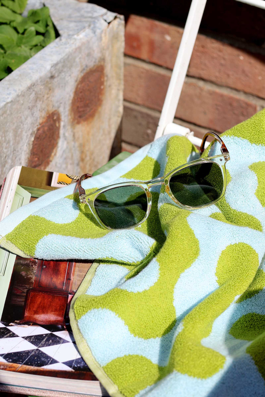 Green sunglasses on a colorful towel with a wooden surface and plant in the background
