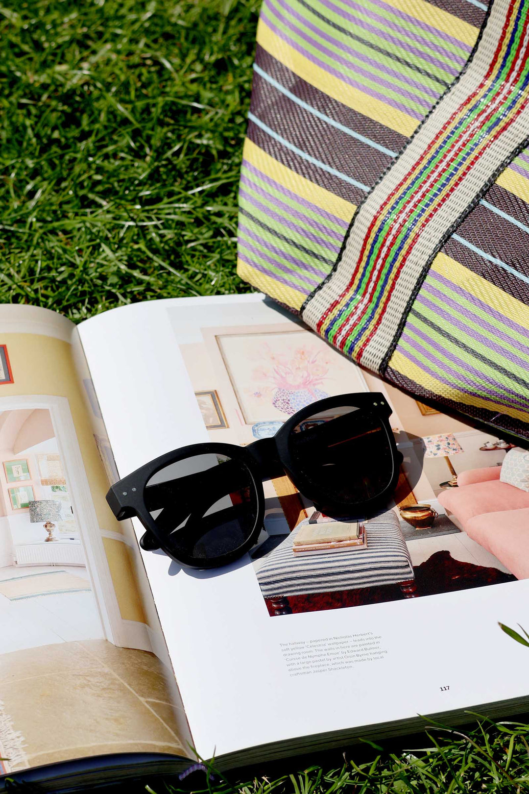 Sunglasses on an open magazine with a colorful striped bag on grass