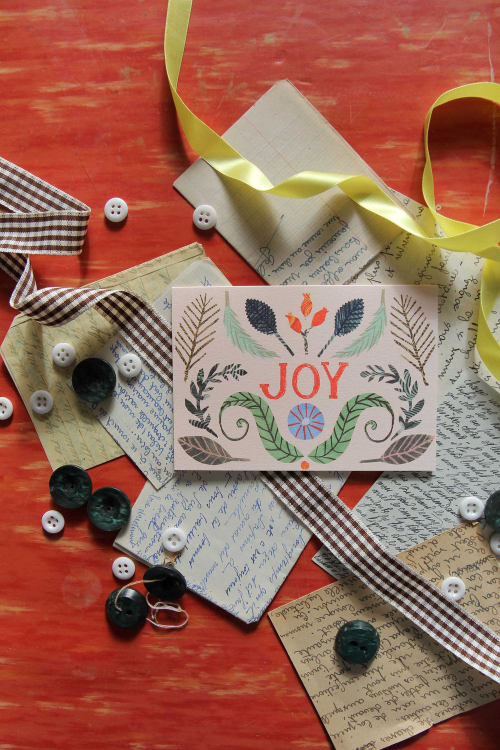 Card with 'JOY' text surrounded by ribbons, buttons, and paper on a red surface