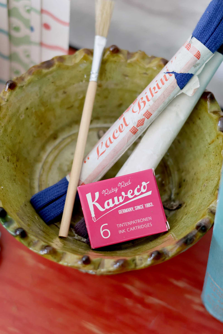 Kaweco ink cartridges with a wooden stick in a green bowl on a red surface