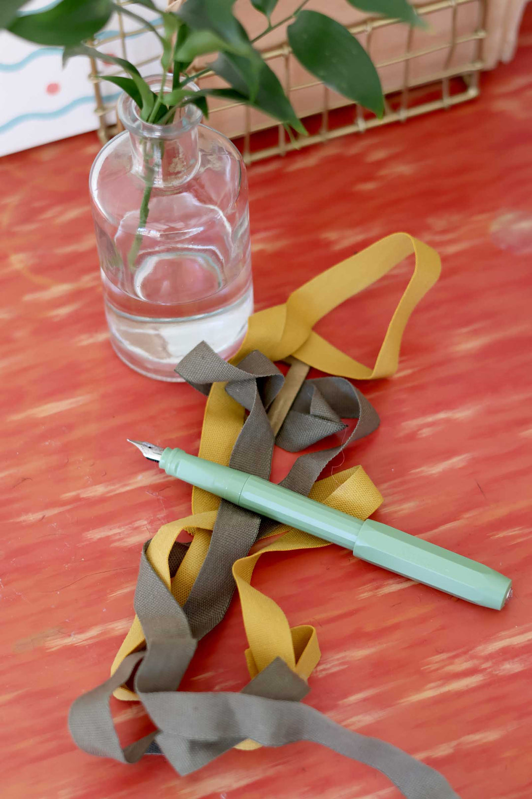 Clear glass vase with water and green plant on a red surface with ribbons and a pen.