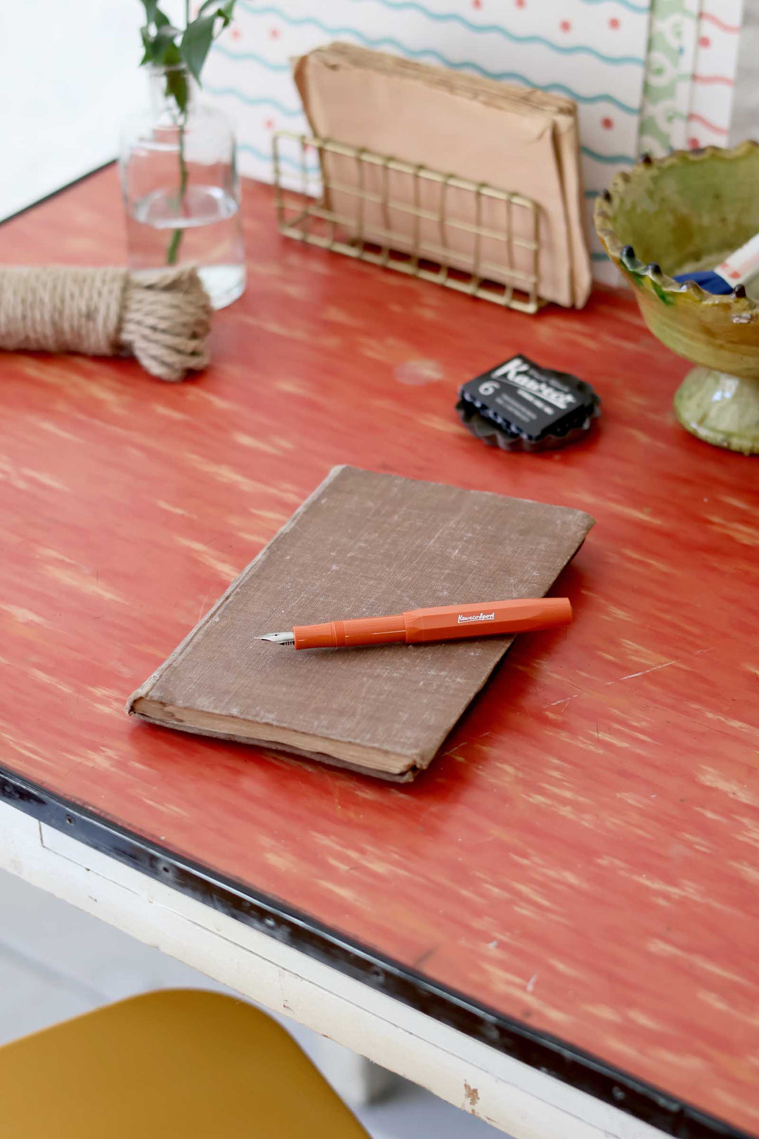 Notebook with a pen on a red desk with a chair and various items in the background
