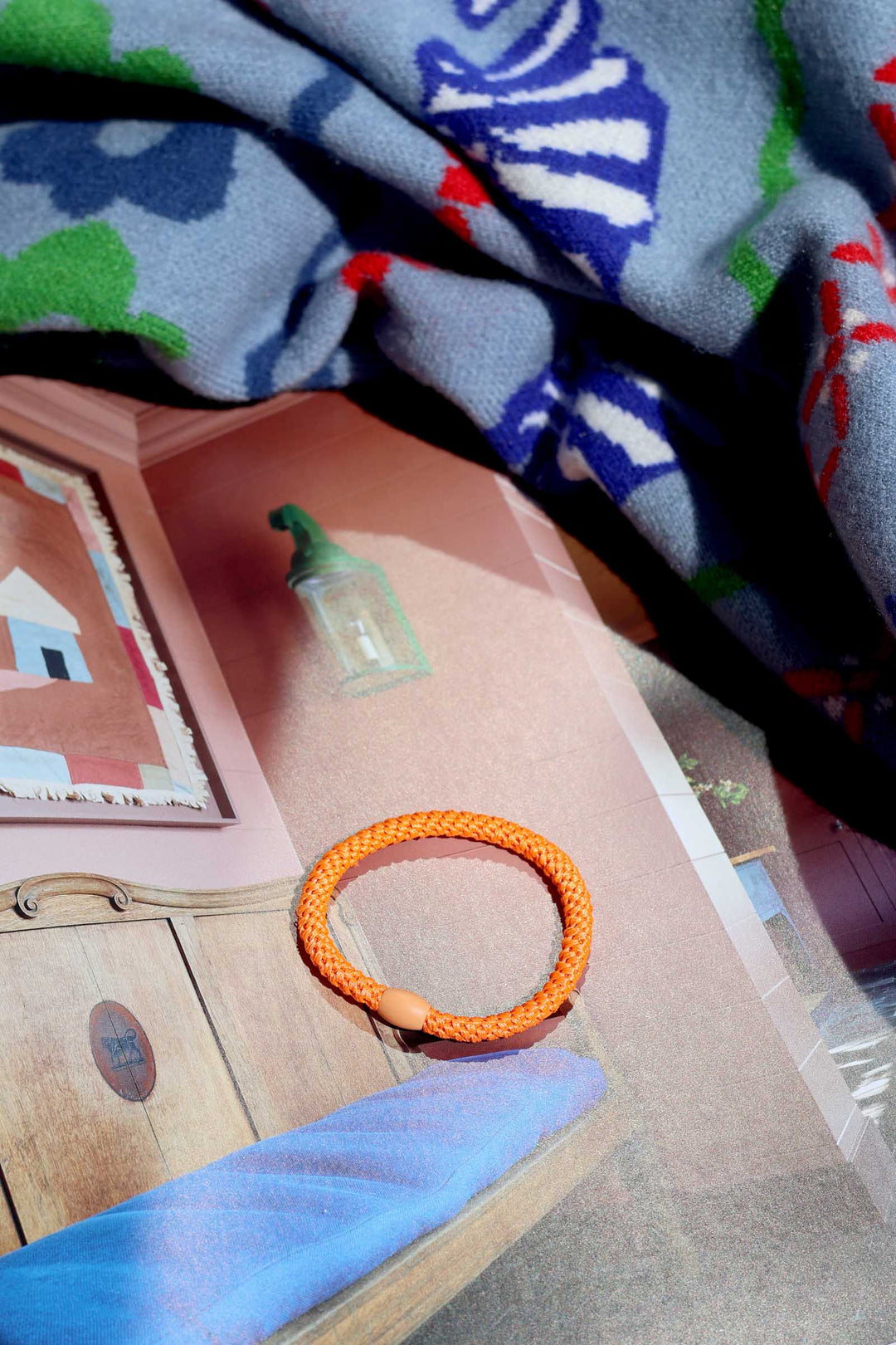 Orange bracelet on a pink surface with a colorful blanket in the background