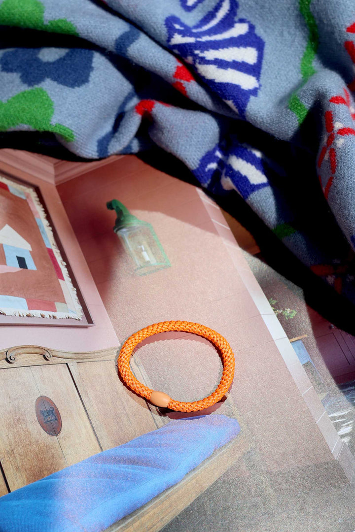 Orange bracelet on a pink surface with a colorful blanket in the background