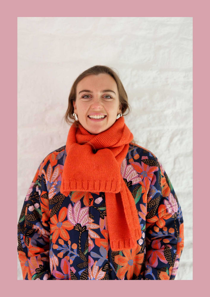 Person wearing a colorful floral-patterned sweater and an orange scarf against a textured white background.