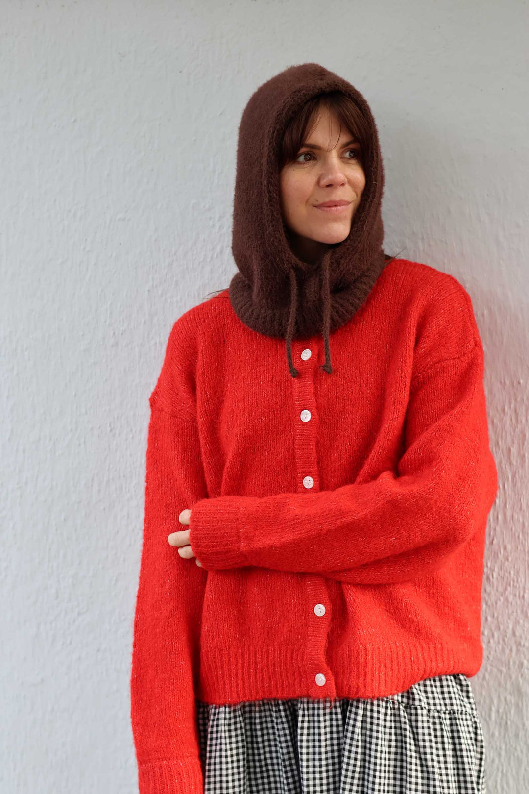 A woman wearing a bright red knit cardigan with a brown hood and a black-and-white gingham skirt, standing against a light gray textured wall.