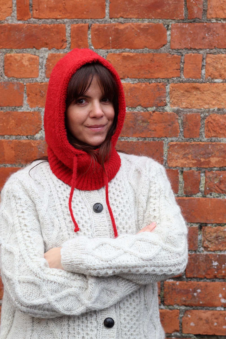 A woman wearing a bright red knit hooded cowligan sweater over a cream cable-knit cardigan, standing with arms crossed in front of a red brick wall.