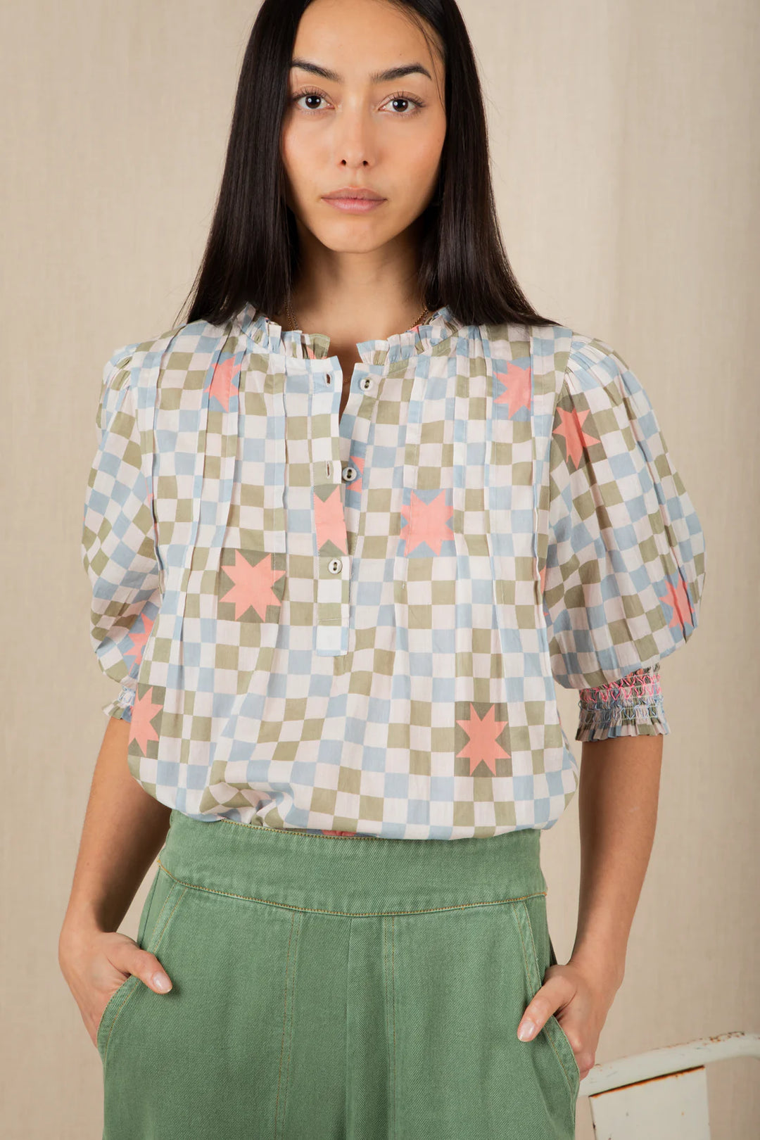 Woman wearing a checkered blouse with star patterns and green pants against a beige background