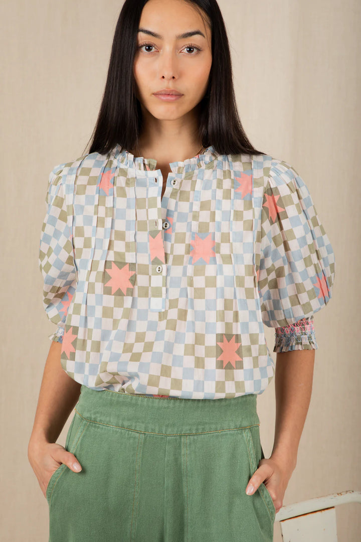 Woman wearing a checkered blouse with star patterns and green pants against a beige background