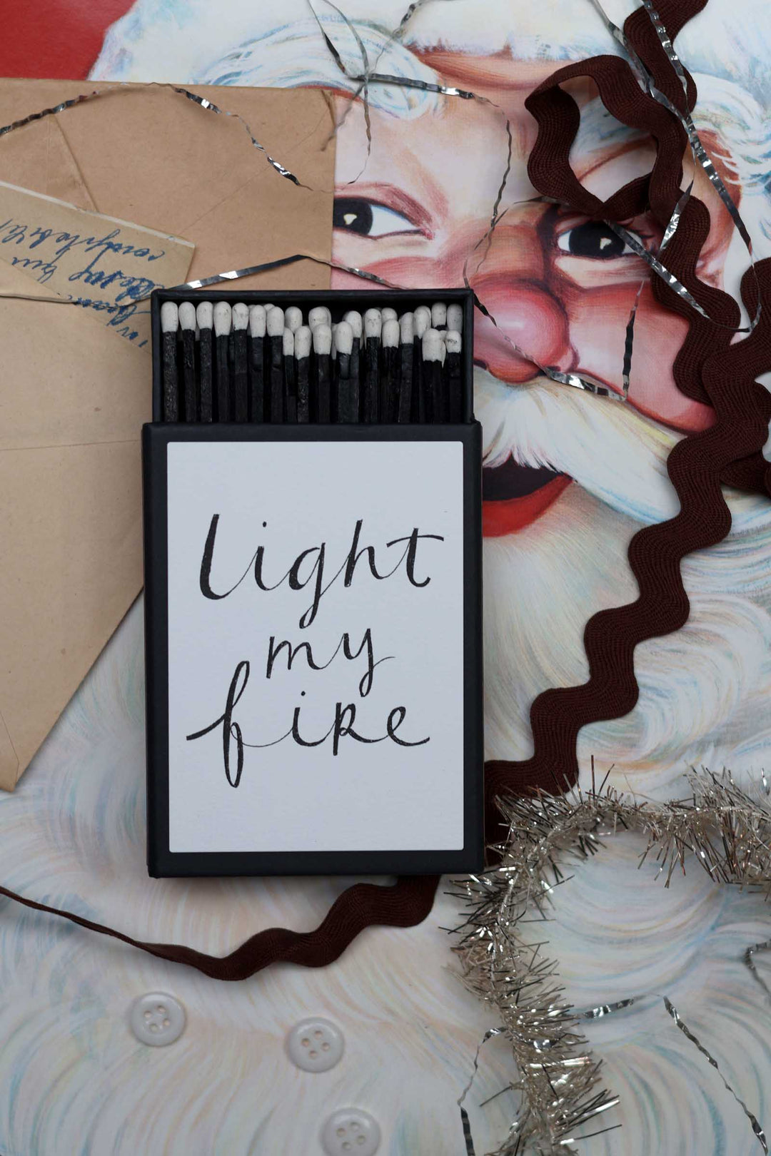 A black matchbox with a white insert reading 'light my fire' in black script, placed on top of a Santa face print with brown and silver tinsel and a kraft envelope around it.