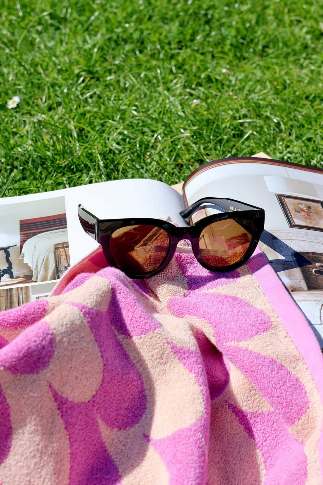 Sunglasses on a pink towel with a magazine underneath, on a grassy background