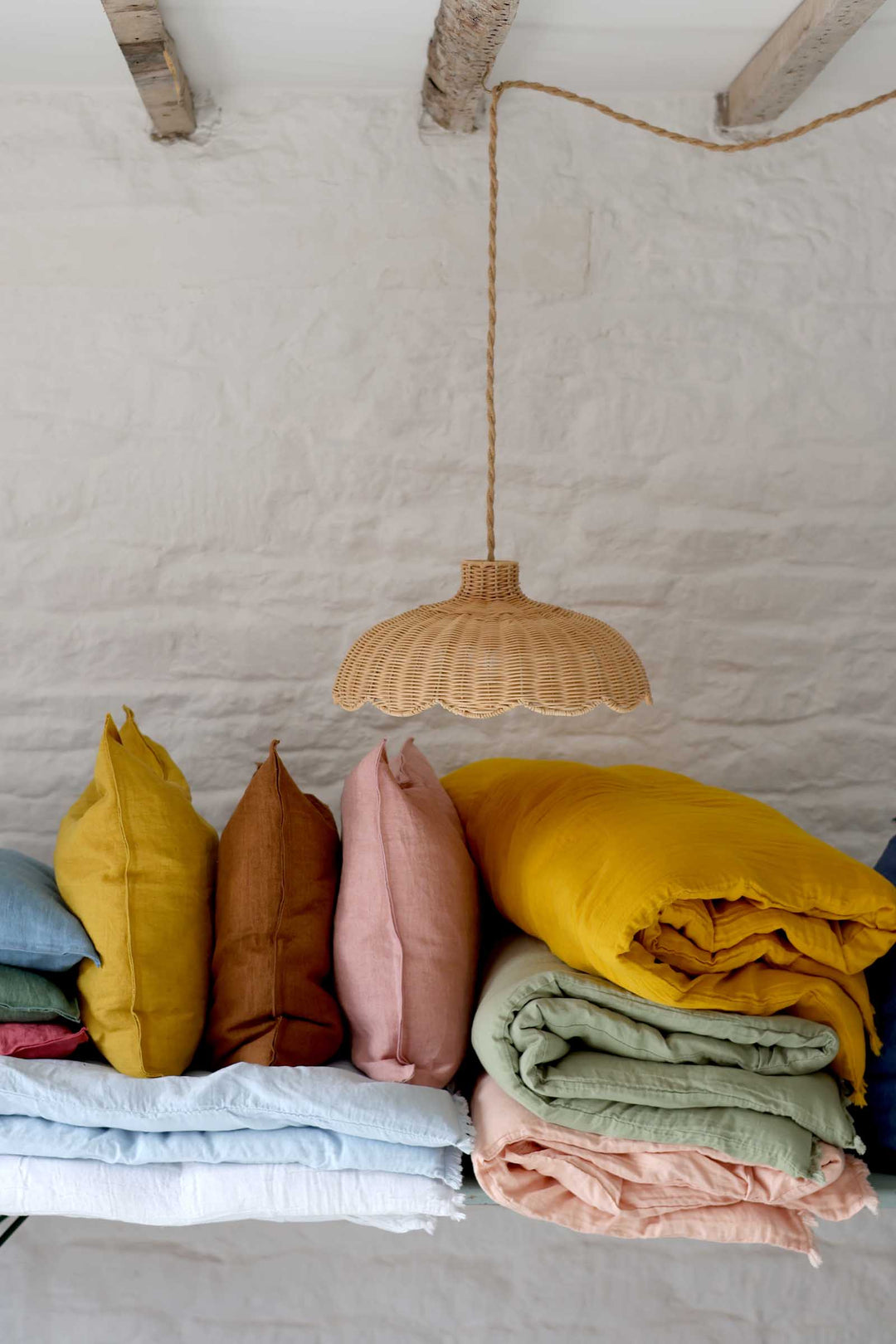 Colorful pillows and blankets under a woven pendant light against a white wall.