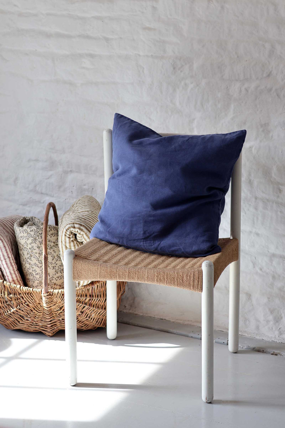 A navy blue linen throw pillow resting on a white-framed chair with a woven seat, set against a white brick wall with a wicker basket nearby.