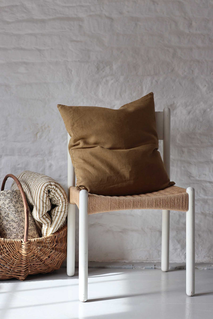 A brown linen throw pillow resting on a white wooden chair with a woven seat, set against a white brick wall with a wicker basket and folded blankets nearby.