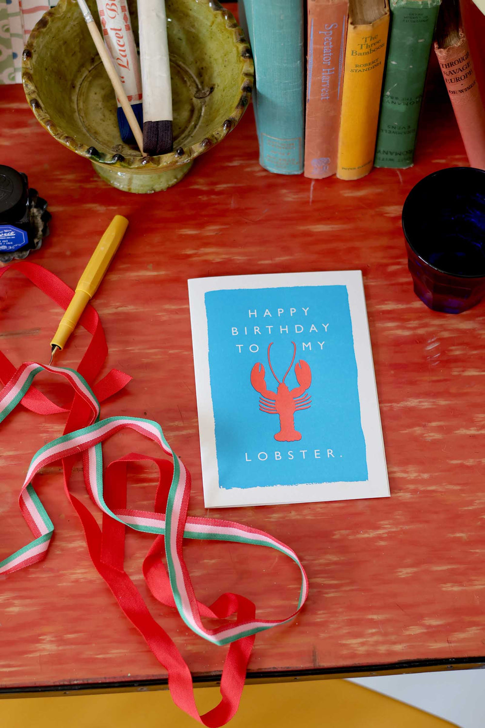 Birthday card with lobster illustration on a red surface with ribbons and stationery items.