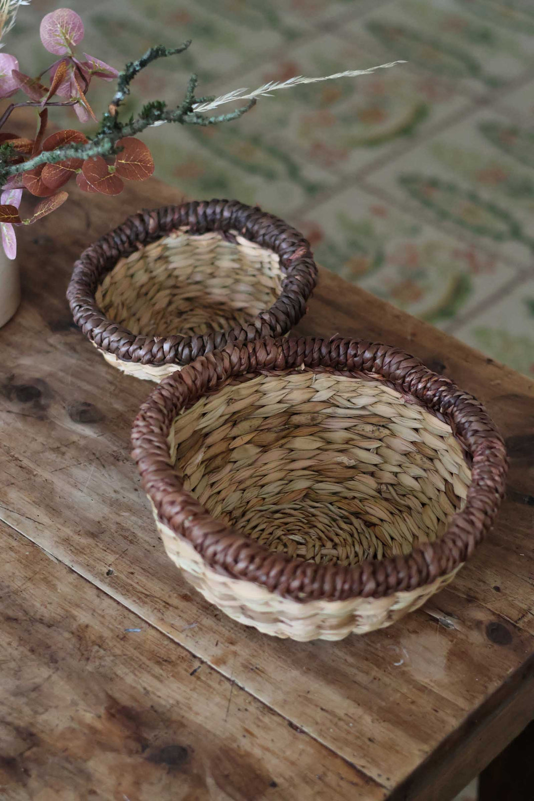 Two round woven baskets with dark brown braided rims and natural straw bodies, displayed on a rustic wooden table.