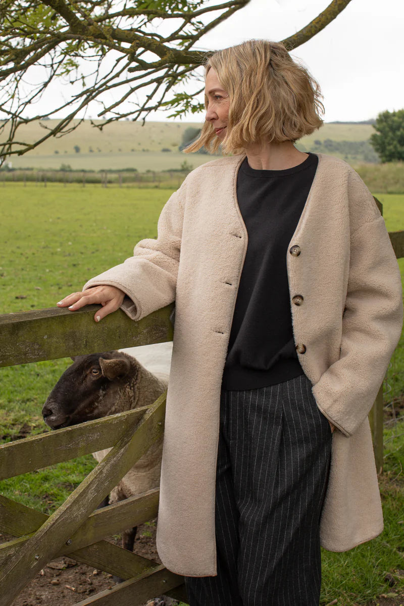 Woman in a beige coat standing next to a wooden gate with a sheep in a field.