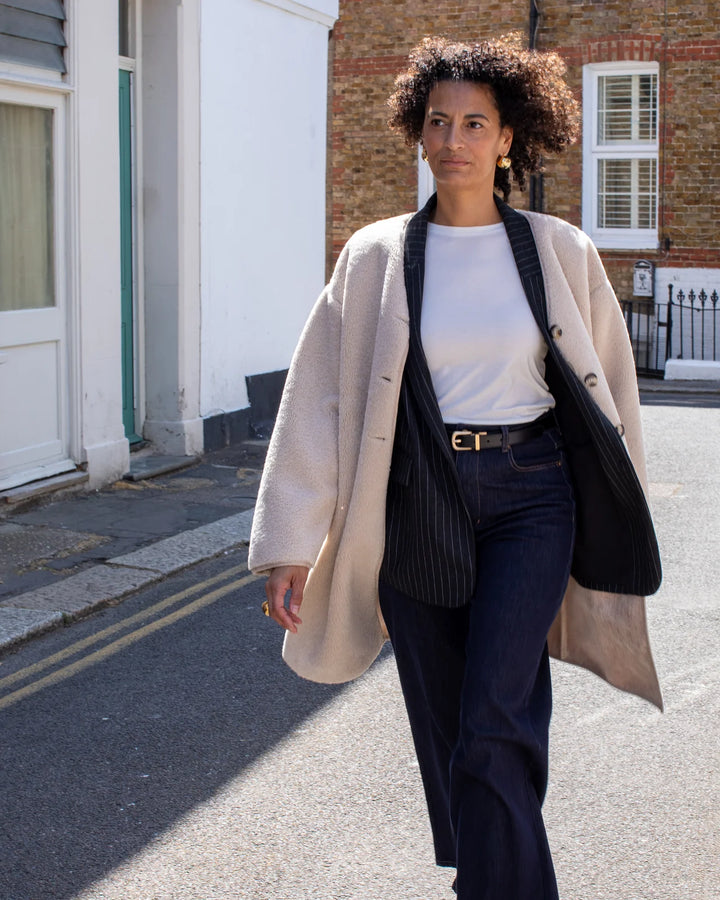 Woman walking on a street wearing a beige coat, white shirt, and dark pants.
