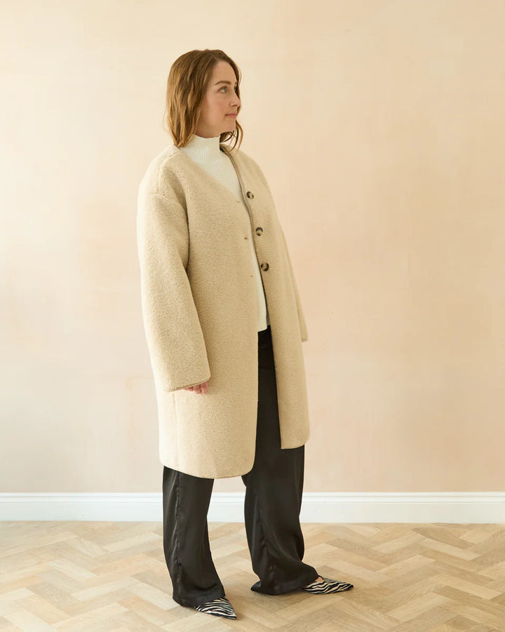 Woman wearing a beige coat standing against a plain wall.