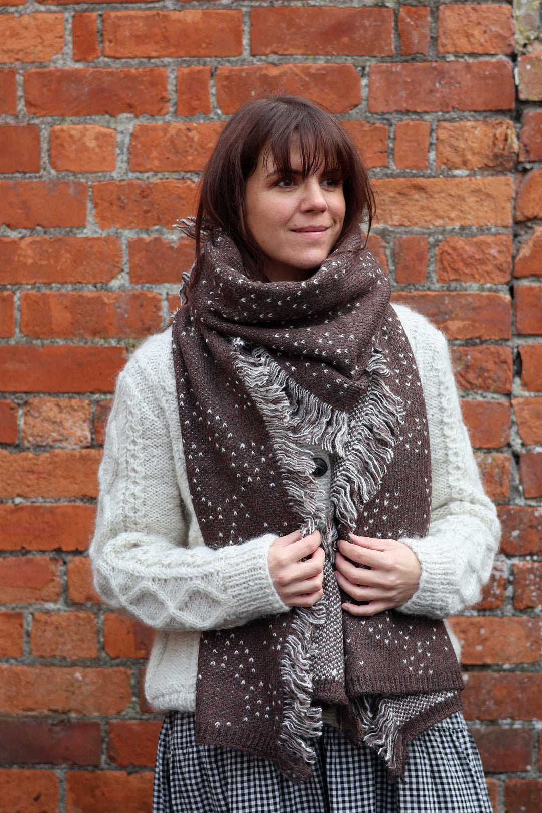 A woman wearing a brown knit scarf with white speckles and fringe, styled over a cream cable-knit sweater and a black-and-white gingham skirt, standing against a red brick wall.