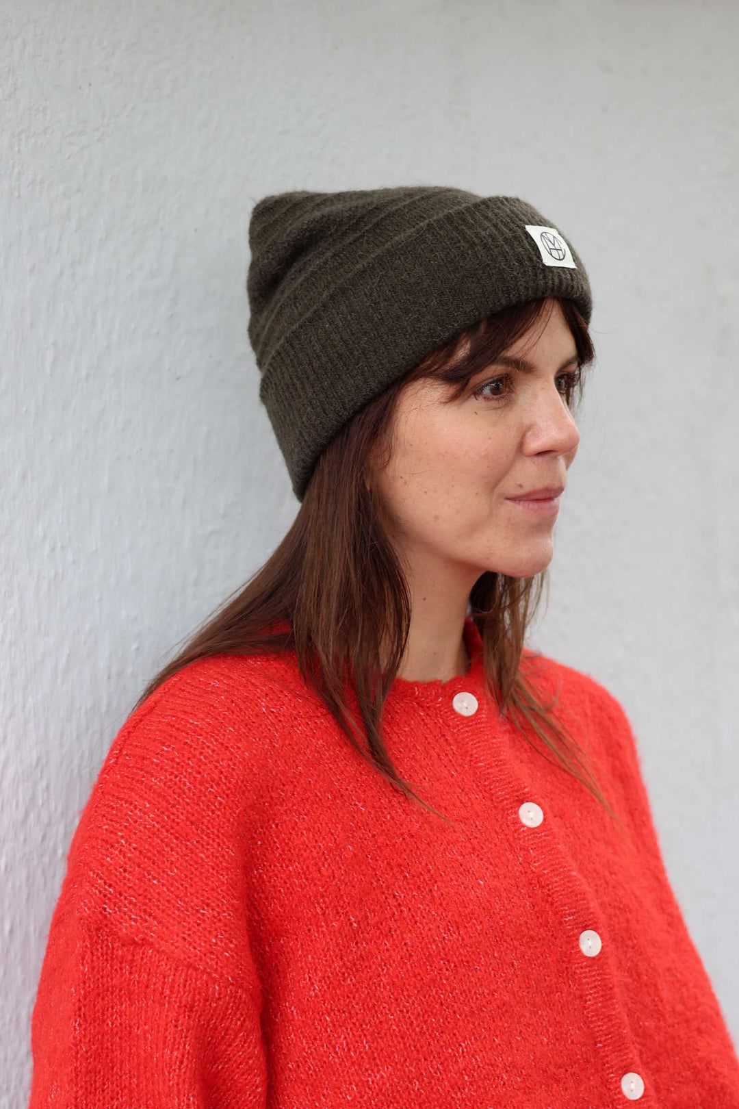 A woman wearing a dark olive green knit beanie with a small white square patch and a bright red knit cardigan, standing against a light gray textured wall.