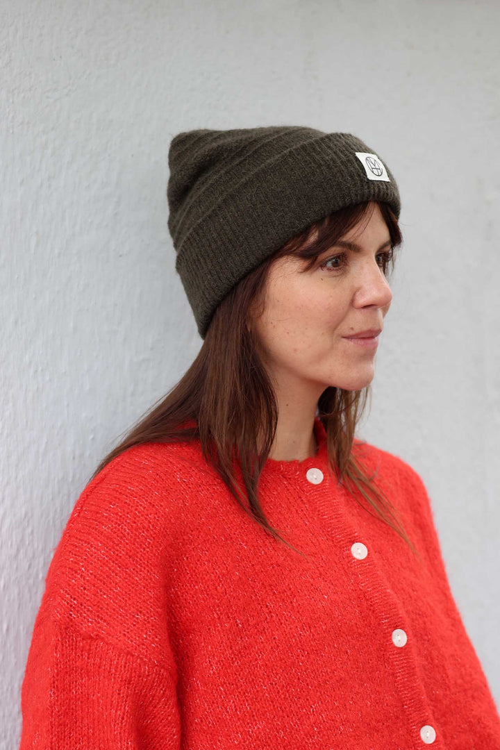 A woman wearing a dark olive green knit beanie with a small white square patch and a bright red knit cardigan, standing against a light gray textured wall.