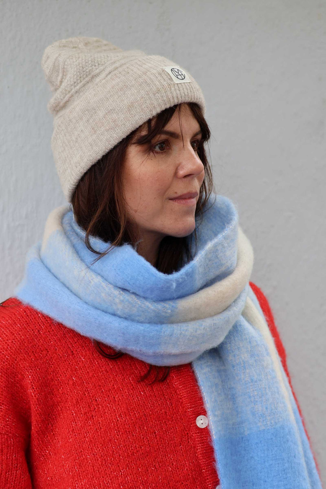 A woman wearing a light beige knit beanie, a thick light blue and cream knit scarf, and a red knit cardigan, photographed against a plain light gray wall.