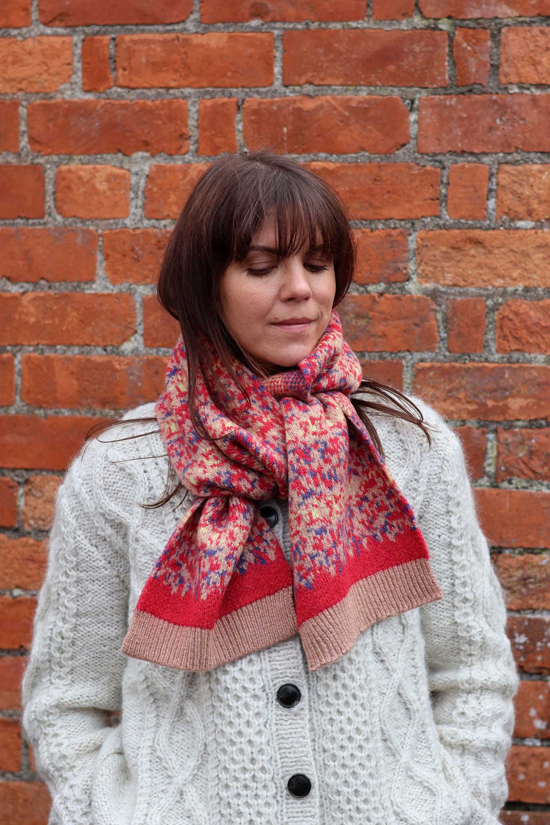 A woman wearing a cream cable-knit cardigan and a red, pink, and blue patterned knit scarf, standing in front of a red brick wall.